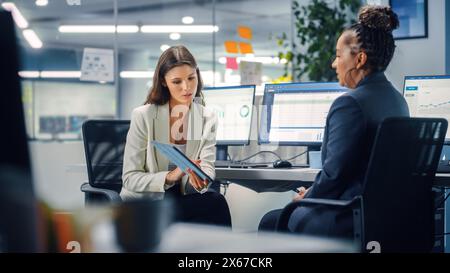 Zwei weibliche Kollegen sprechen liebevoll miteinander, lächeln, während sie in verschiedenen modernen Business-Office-Büros Arbeit am Laptop zeigen. Erfahrener Manager und junger Mitarbeiter diskutieren ein unterhaltsames analytisches Projekt. Stockfoto