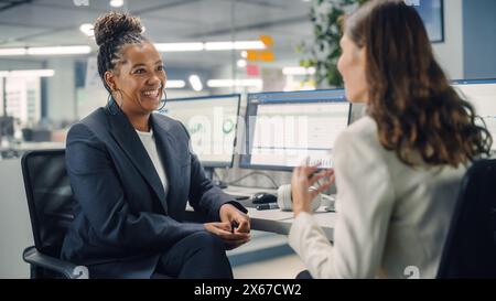 Zwei weibliche Kollegen sprechen liebevoll miteinander, lachen und lächeln, während sie an Computern in verschiedenen modernen Geschäftsbüros arbeiten. Erfahrener Manager und junger Mitarbeiter diskutieren ein unterhaltsames analytisches Projekt. Stockfoto