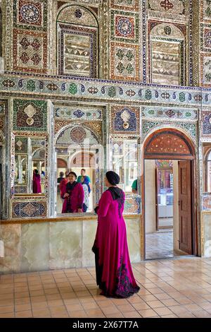 Die iranische Frau in einem schicken Kleid schaut in den Spiegel des Zinat Al-Molk Historical House, einer Residenz aus der Qajar-Zeit aus dem 19. Jahrhundert. Shiraz, Iran. Stockfoto