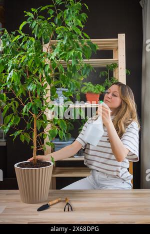 Eine konzentrierte Frau bewässert eine große, belaubte Innenpflanze mit einer weißen Gießkanne in ihrem städtischen Garten, mit Gartengeräten auf dem Tisch. Stockfoto