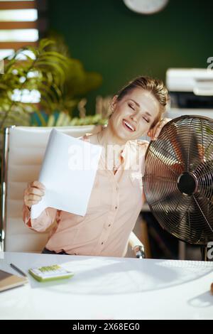 Nachhaltiger Arbeitsplatz. Glückliche moderne 40-jährige Buchhalterin im modernen grünen Büro mit Dokumenten und elektrischem Ventilator. Stockfoto