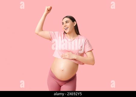 Glückliche schwangere Frau, die Muskeln vor rosa Hintergrund zeigt Stockfoto