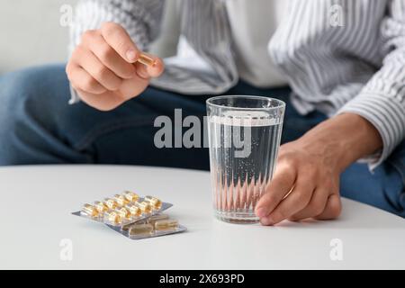 Hübscher junger Mann mit Blisterpackungen Fischöl Kapseln und Wasser, der zu Hause auf dem Sofa sitzt, Nahaufnahme Stockfoto