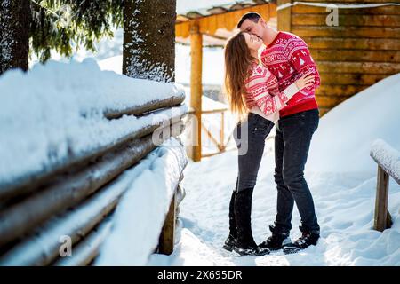 Verlobungssitzung, Liebesgeschichte eines jungen heterosexuellen Paares, das sich in weihnachtlich roten Pullis und Jeans in verschneiten Landschaften in der Nähe eines wo umarmt und küsst Stockfoto