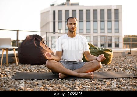 Mann meditiert in Lotusposition, während er auf der Matte auf dem Dach des modernen Hauses sitzt. Stockfoto