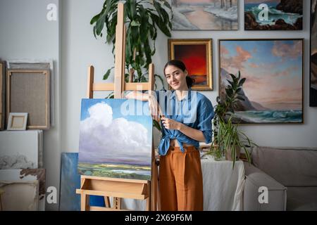 Selbstbewusste Künstlerin, die Ölgemälde zeigt. Malerin, die mit einem versicherten Lächeln in die Kamera blickt Stockfoto