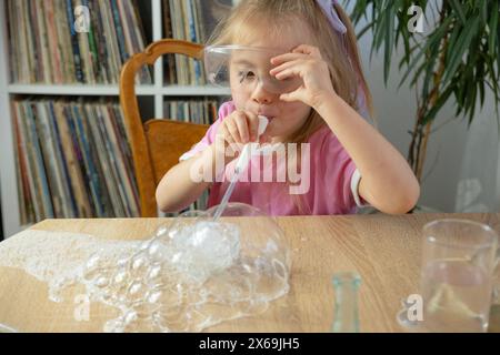 Das kleine Mädchen bläst Blasen durch Plastikstroh aus Seifenschaum, lustige Spiele für Kinder, selbstgebaute Seifenblasen, macht Experimente Stockfoto