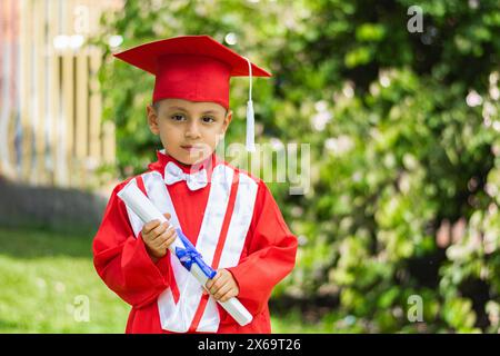 Porträt eines kleinen lateinjungen im roten Abschlusskleid, posiert während des Diploms im Park Stockfoto