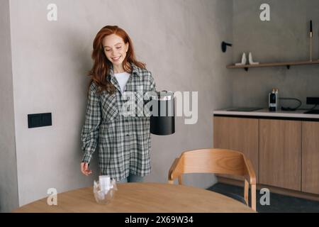 Porträt einer glücklichen lächelnden jungen Frau, die Kaffee mit Tropfkaffeesack in einer transparenten Glasschale macht, die am Holztisch steht. Vorbereitung frisch verarbeiten Stockfoto