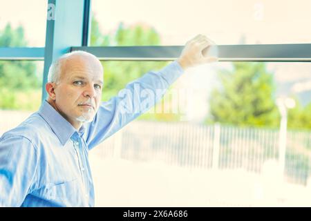 Nachdenklicher älterer Mann in blauem Hemd reflektiert seine Zukunft und Familie, blickt aus einem großen Fenster in einem sonnendurchfluteten Raum, denkt über den Ruhestand und Hi Stockfoto