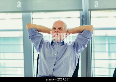 Bequem sitzend nimmt der Mann einen Moment der Ruhe, umgeben von der lichtdurchfluteten Atmosphäre seines Arbeitsplatzes Stockfoto