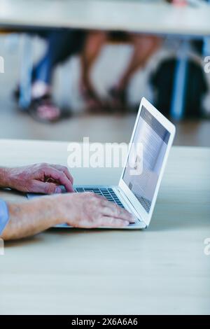 Die Hände eines älteren Mannes tippen auf einem Laptop in einem hellen Raum, auf dem verschwommenen Hintergrund Beine und Füße zu sehen sind. Der Mann trägt ein blaues Hemd und konzentriert sich auf den Stockfoto