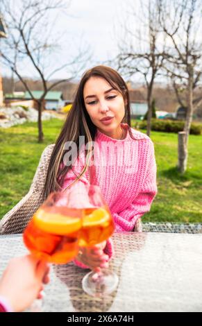 Porträt einer jungen russischen Frau, die Cocktail-aperol-Spritz trinkt, draußen in rosa Pullover sitzt Stockfoto