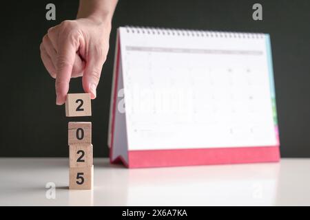 Hand auf 2025 Holzwürfel mit Kalender im Hintergrund. Neues Jahr, Neubeginn. Kopierbereich. Stockfoto