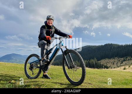 Radfahrer, der draußen mit dem Elektrofahrrad unterwegs ist. Porträt eines lächelnden männlichen Touristen, der auf einem grasbewachsenen Hügel ruht, die wunderschöne Berglandschaft genießt und Helm trägt. Konzept der aktiven Freizeit. Stockfoto