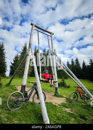 Person, die auf einer großen Holzschaukel auf einem Hügel oder Berg sitzt, geeignet für Freizeitaktivitäten im Freien. Zwei E-Mountainbikes, die sich gegen den Schaukelrahmen lehnen. Stockfoto