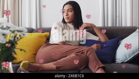 Bild von Herzsymbolen mit Blumen über birassischer Frau, die auf Sofa sitzt Stockfoto