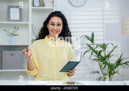 Eine lächelnde hispanische Frau, die ein Online-Tutorial mit einem Schüler durchführt, Gesten verwendet und ein Notizbuch in einer hellen Büroumgebung hält. Stockfoto