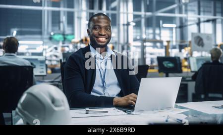 Porträt eines glücklichen jungen afroamerikanischen Ingenieurs, der an einem Laptop-Computer in einem Büro in der Automontageanlage arbeitet. Industriespezialist, der an Fahrzeugteilen in der technischen Entwicklungsanlage arbeitet. Stockfoto