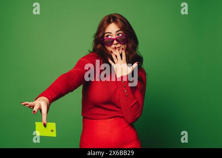 Eine junge Frau mit Sonnenbrille hält eine grüne Kreditkarte in einem lebendigen Studio. Sie zeigt einen modernen und modischen Lebensstil und macht nahtlos Elektro Stockfoto