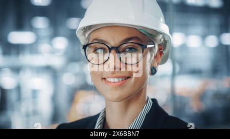 Porträt einer glücklichen, lächelnden jungen, schönen Ingenieurin mit Brille und weißem Schutzhelm im Büro in der Autowerkstatt. Industriespezialist, der an Fahrzeugdesign in modernen Anlagen arbeitet. Stockfoto