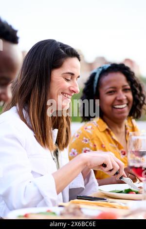 Seitenansicht einer lächelnden kaukasischen Frau, die Essen isst und mit Freunden auf dem Dach am Tisch sitzt. Stockfoto