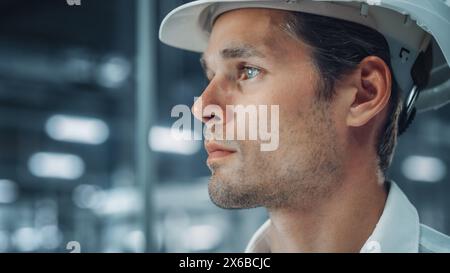 Porträt eines jungen gutaussehenden, selbstbewussten Ingenieurs mit weißem Schutzhelm im Büro in der Autowerkstatt. Industriespezialist, der an der Fahrzeugproduktion in der modernen Fabrik arbeitet. Seitenprofil, Flächenansicht. Stockfoto