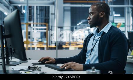 Autowerkstatt: Porträt eines selbstbewussten männlichen Chefingenieurs, der am Desktop-Computer arbeitet. Professioneller Techniker in der Herstellung von automatisierten Roboterarmen in der Fertigung von High-Tech-Elektrofahrzeugen Stockfoto