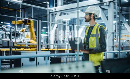 Autofabrik: Männlicher Kfz-Ingenieur Mit Schutzhelm, Stehend, Mit Laptop. Überwachung, Steuerung, Geräteproduktion. Automatisierte Roboterarm-Montage-Fertigungslinie Für Elektrofahrzeuge Stockfoto