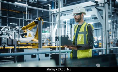 Autofabrik: Männlicher Kfz-Ingenieur Mit Schutzhelm, Stehend, Mit Laptop. Überwachung, Steuerung, Geräteproduktion. Automatisierte Roboterarm-Montage-Fertigungslinie Für Elektrofahrzeuge Stockfoto