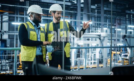 Autofabrik: Projektmanager und Automobilingenieur in Hard hat, Talking, Using Laptop. Überwachung, Steuerung, Geräteproduktion. Automatisierte Roboterarm-Montage-Fertigungslinie Für Elektrofahrzeuge Stockfoto