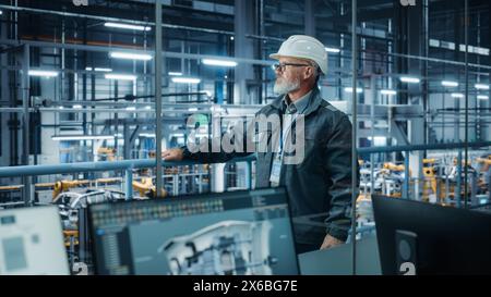 Autofabrik: Selbstbewusster männlicher Kfz-Ingenieur, der einen Schutzhelm trägt, Steh- und Überwachungsausrüstung Produktionslinie. Automatisierte Roboterarm-Montage-Fertigungslinie Für Elektrofahrzeuge Stockfoto