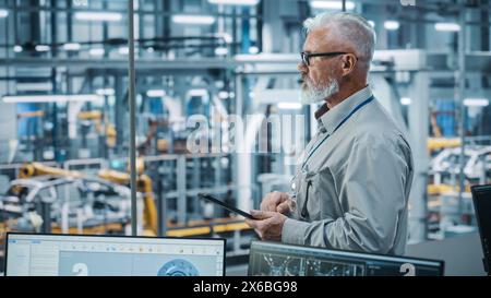Autowerkstatt: Porträt eines leitenden weißen männlichen Chefingenieurs mit Tablet-Computer in automatisierten Roboterarmmontagestraßen zur Herstellung von High-Tech-Elektrofahrzeugen. Seitenansicht Stockfoto
