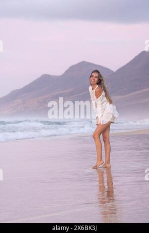 Die junge Frau lächelt, während sie barfuß an einem Sandstrand spaziert, mit sanften Wellen und pastellfarbenen Sonnenuntergängen über fernen Bergen, die eine ruhige Kulisse schaffen Stockfoto