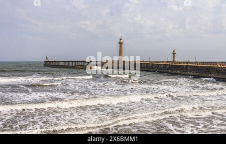 Ein Pier mit einem Leuchtturm erstreckt sich in ein stürmisches Meer, während Wellen mit weißem Schaum an die Küste dröhnen. Ein zweiter Leuchtturm ist dahinter. Stockfoto