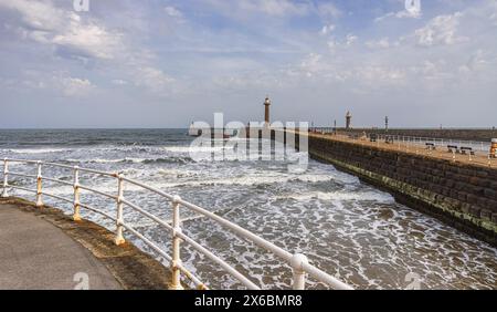 Ein Pier mit einem Leuchtturm erstreckt sich in ein stürmisches Meer, während Wellen mit weißem Schaum an die Küste dröhnen. Ein zweiter Leuchtturm ist dahinter. Stockfoto