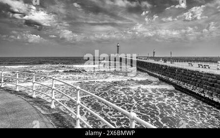 Ein Pier mit einem Leuchtturm erstreckt sich in ein stürmisches Meer, während Wellen mit weißem Schaum an die Küste dröhnen. Ein zweiter Leuchtturm ist dahinter. Stockfoto