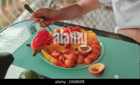 Eine Person sitzt an einem Tisch und isst geschickt einen Teller mit verschiedenen Früchten. Die Szene zeigt eine Vielzahl von Früchten, die in kleinere Stücke geschnitten werden, exotische Früchte Stockfoto