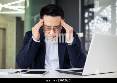 Asiatischer reifer Mann in einem Business-Anzug, der Stress und Kopfschmerzen erlebt, während er im Büro an einem Laptop arbeitet. Stockfoto
