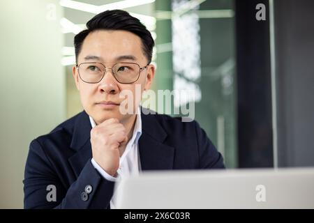 Asiatischer reifer Mann in Anzug und Brille, der an einem Schreibtisch denkt und arbeitet. Geschäftskonzept. Stockfoto