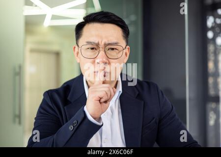 Asiatischer reifer Mann in Geschäftskleidung, der eine scheußende Geste macht und Stille und Vertraulichkeit in einem Büro betont. Stockfoto