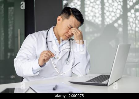 Asiatischer reifer Arzt sieht gestresst aus, während er mit Laptop und Brille am Schreibtisch sitzt und Stress und Ermüdung vermittelt. Stockfoto