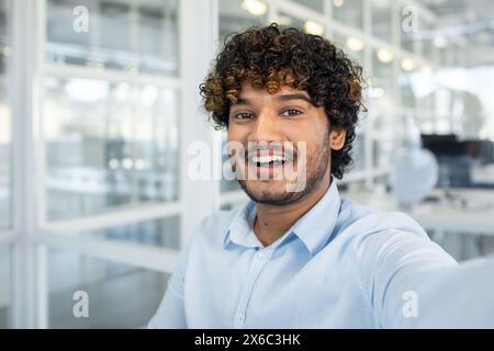 Fröhlicher junger Mann, der ein Selfie in einem hellen, modernen Bürobereich macht, der Freude und eine professionelle Atmosphäre zeigt. Stockfoto