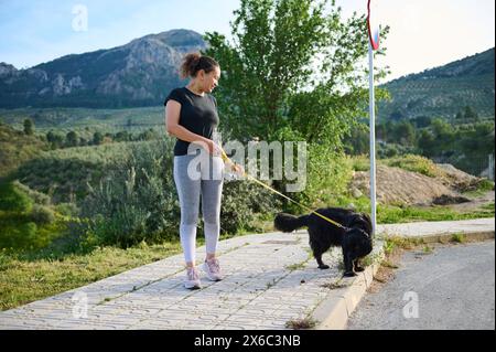 Porträt einer glücklichen Frau, die ihren Hund an der Leine in der Natur spaziert. Multiethnische Spaziergänge mit ihrem Pedigree reinrassig schwarz c Stockfoto