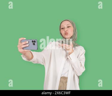 Porträt eines kaukasischen jungen Mädchens im Hijab, der Luftkuss bläst. Halten Sie das Smartphone, um Selfie zu machen. Videoaufzeichnung, Online-Anruf. Schöne muslimische Frau. Stockfoto