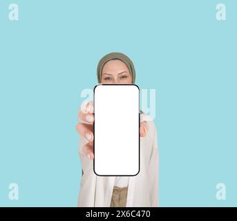 Kaukasische muslimische Frau im Hijab, die Telefonvorstellungen zeigt. Smartphone bedeckt die Hälfte ihres Gesichts. Selektiver Fokus auf Hand. Weißes leeres Display. Stockfoto
