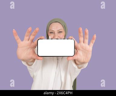 Lächelnde muslimische Frau im Hijab, die ein horizontales Telefon zeigt. Mit zwei Händen halten. Nahaufnahme modernes Smartphone-Modell Stockfoto