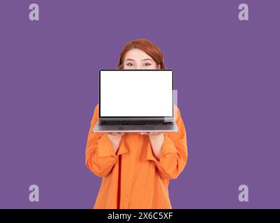 Rotes Haar Frau, die hinter einem modernen Laptop nachschaut. Unerkennbares Mädchen hatte Angst oder schockierte große blaue Augen. Steht über violettem Hintergrund. Stockfoto