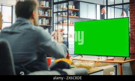 Zu Hause sitzt man auf einer Couch und schaut Green Screen Chroma Key TV. Männer mittleren Alters ausruhen, Watches Show, Sportereignis, Film, On-Demand-Streaming-Service, isst Snacks. Schuss über die Schulter Stockfoto