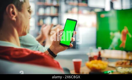 Zu Hause Fußball-Fan hält Green Screen Chroma Key Smartphone, Freunde sehen Fußball-Spiel im Fernsehen, feuern Favourtite Sports Team an, um die Meisterschaft zu gewinnen. Football Club Play. Über der Schulter Stockfoto